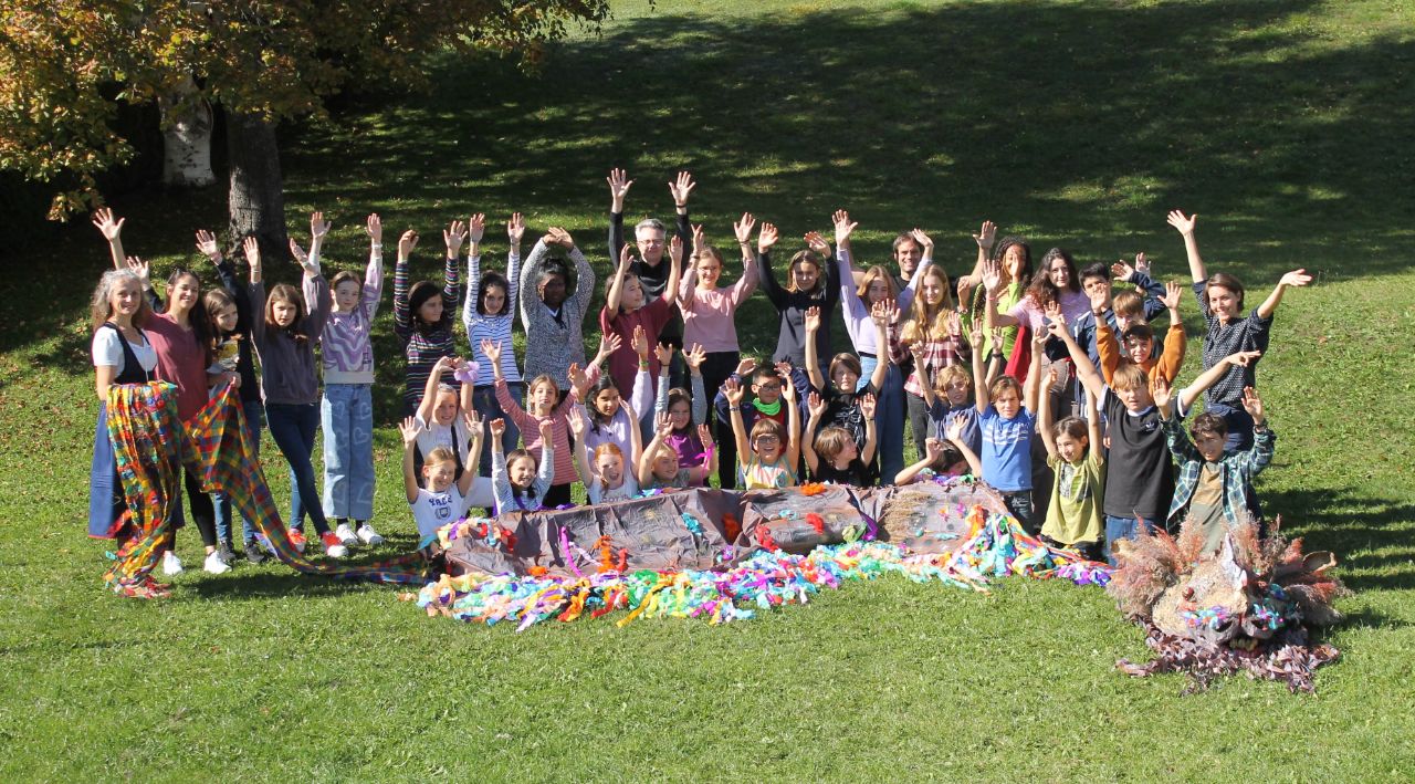 Eine Gruppe von Kindern und Erwachsenen auf einer sonnigen Wiese, vor ihnen liegt ein Drachen aus Papier, sie heben alle die Hände.