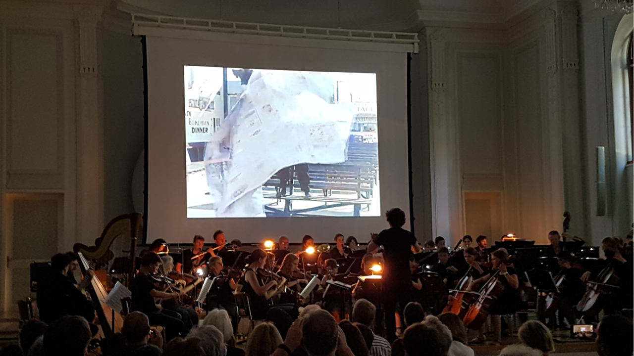 Orchester spielt vor einer Leinwand in einem verdunkelten Saal