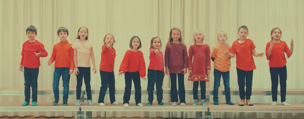 Elf Kinder in rot-schwarzen Kleidern singen und gestikulieren auf der Bühne. 